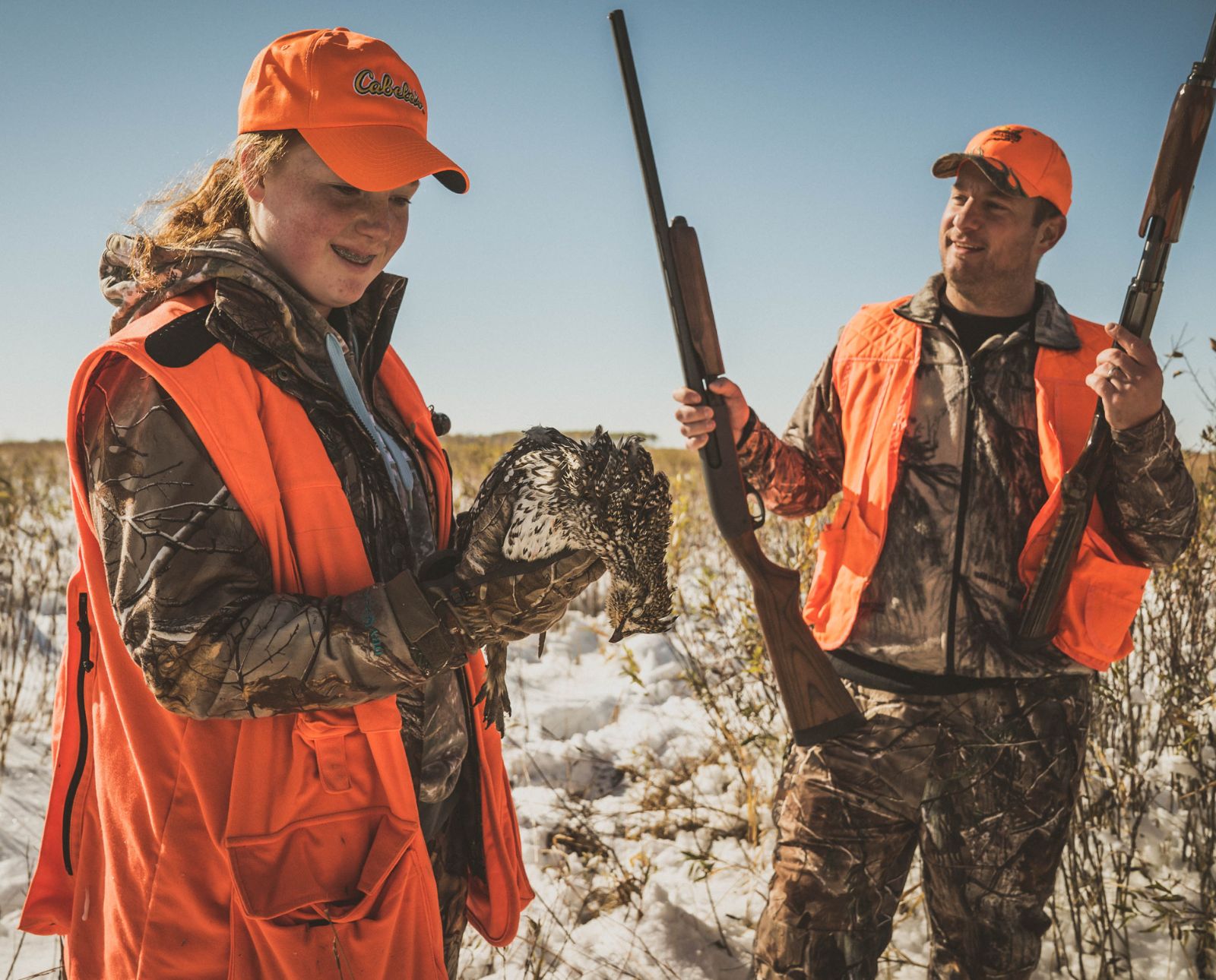 A female upland hunter holds a sharp-tailed grouse while her dad holds two shotguns.
