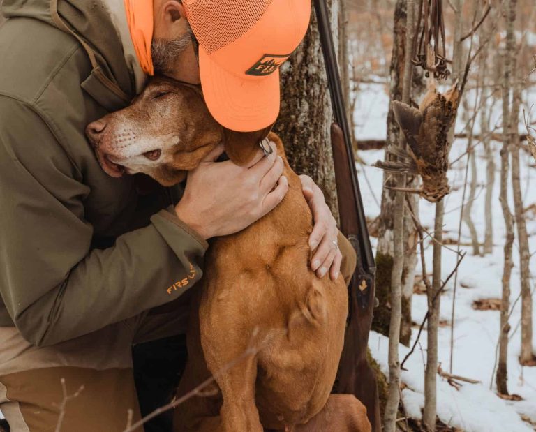 A Vizsla with one front leg hugs his owner in the woods next to a hanging dead American woodcock.