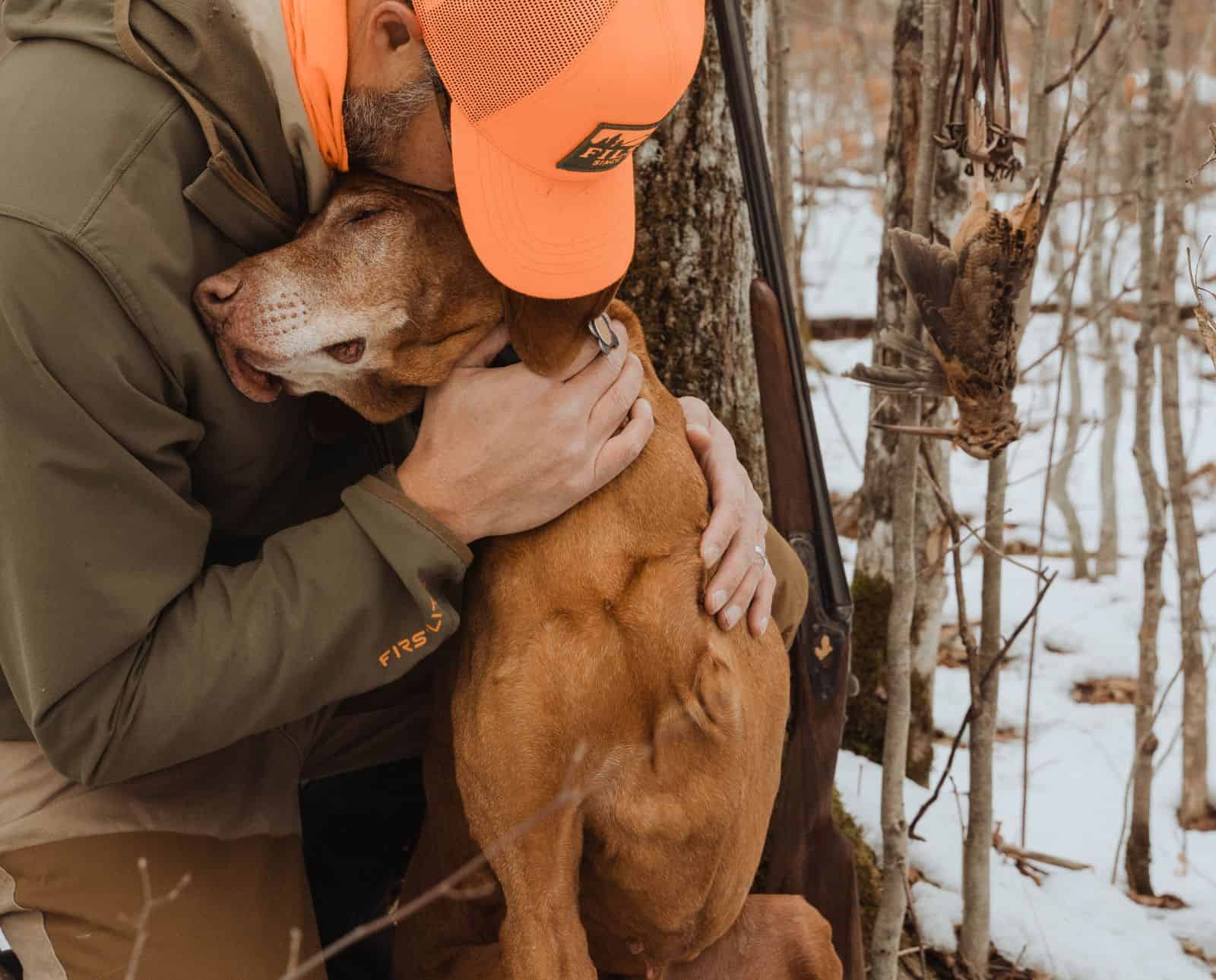 A Vizsla with one front leg hugs his owner in the woods next to a hanging dead American woodcock.