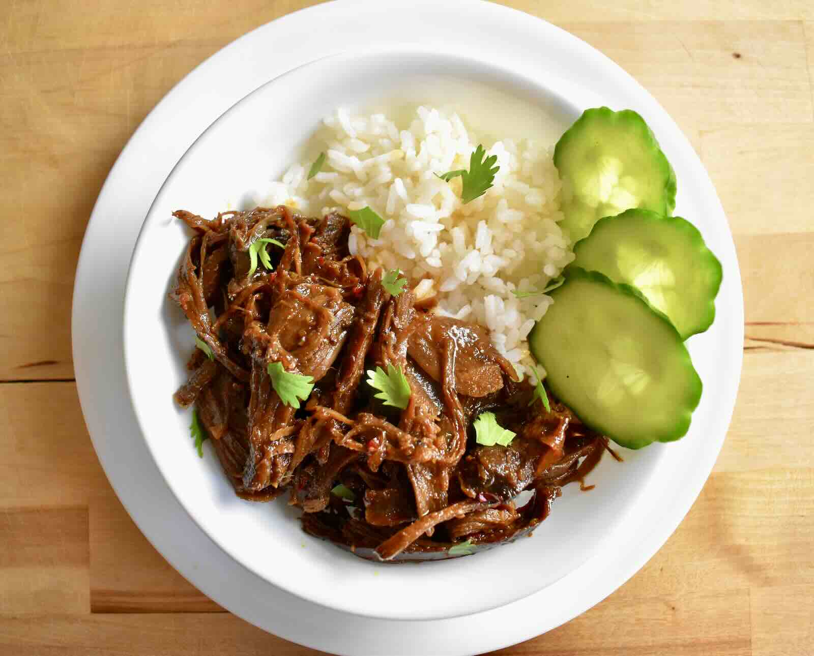Braised and shredded turkey meat is coated in a caramel sauce and served with white rice and cucumber slices in a white bowl