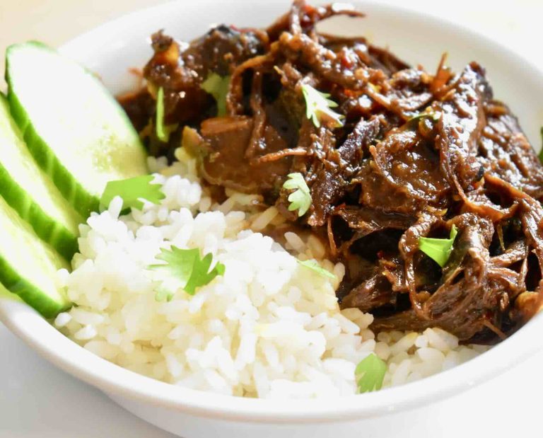 Braised and shredded turkey meat is coated in a caramel sauce and served with white rice and cucumber slices in a white bowl