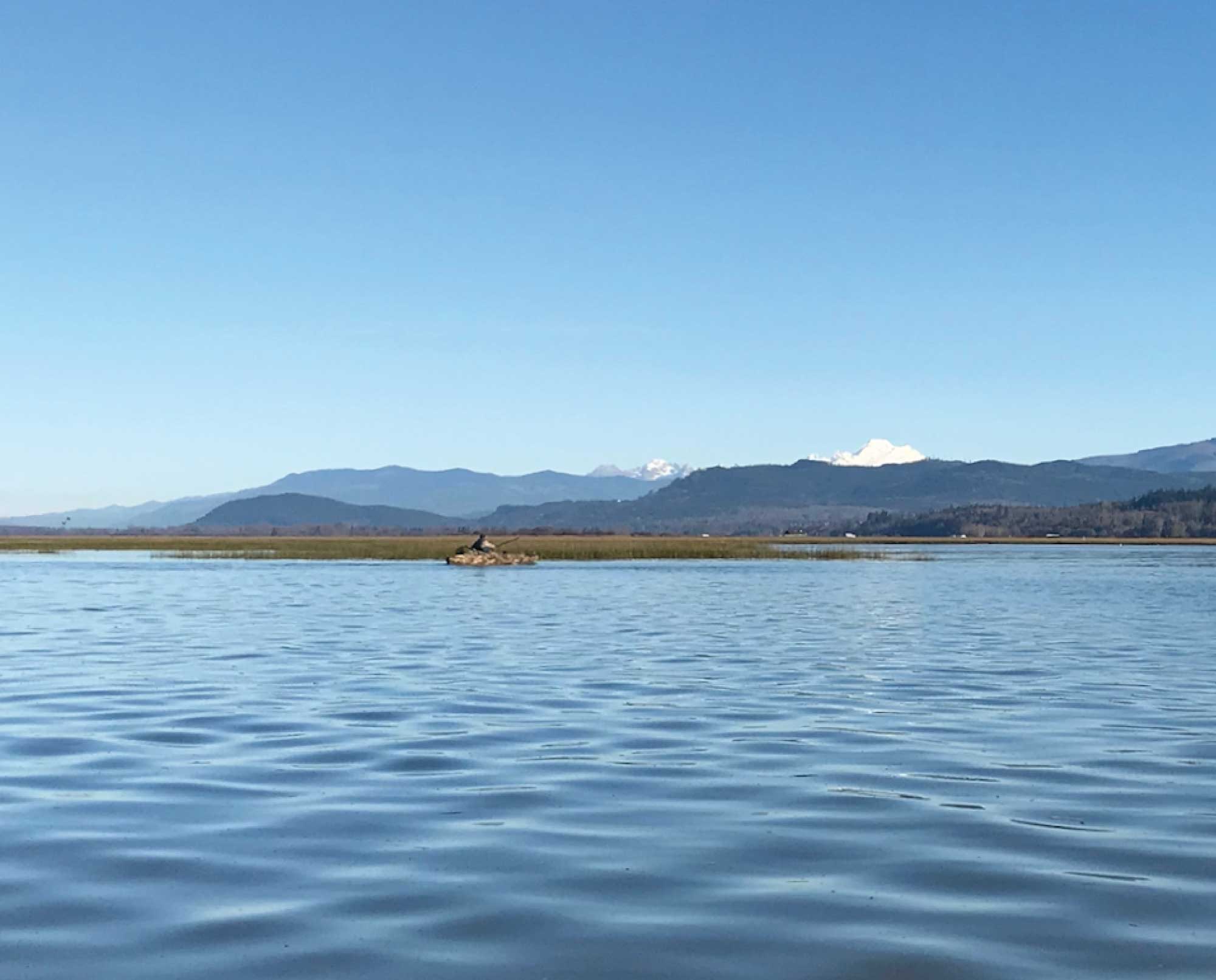 A hunter in a camouflaged kayak paddles in front of a mountain backdrop