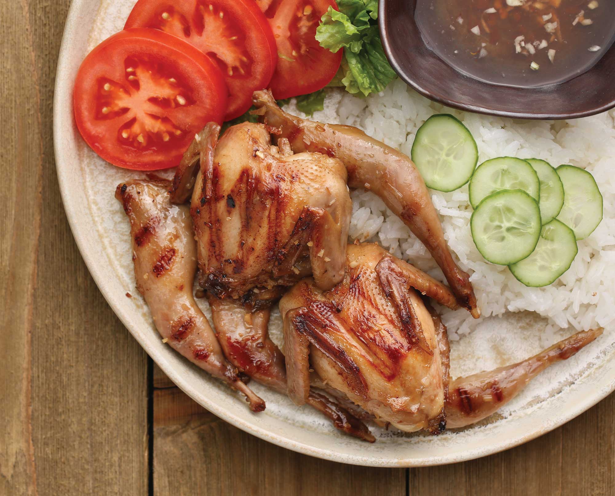 Lemongrass Grilled Quail plated with rice, tomatoes, lettuce, and cucumber in traditional Vietnamese style