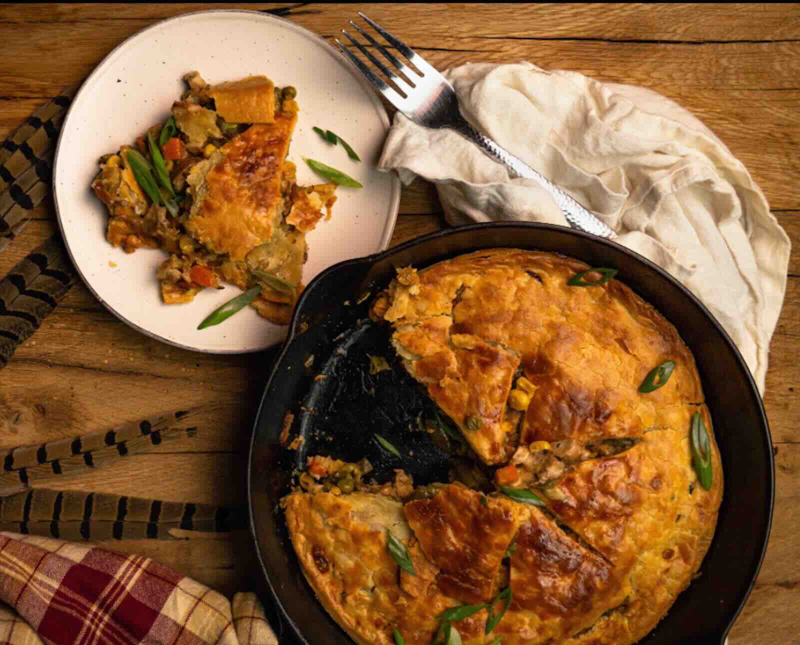 A skillet with a pot pie, one slice removed and served on a white plate, on a wooden background with decorative pheasant feathers