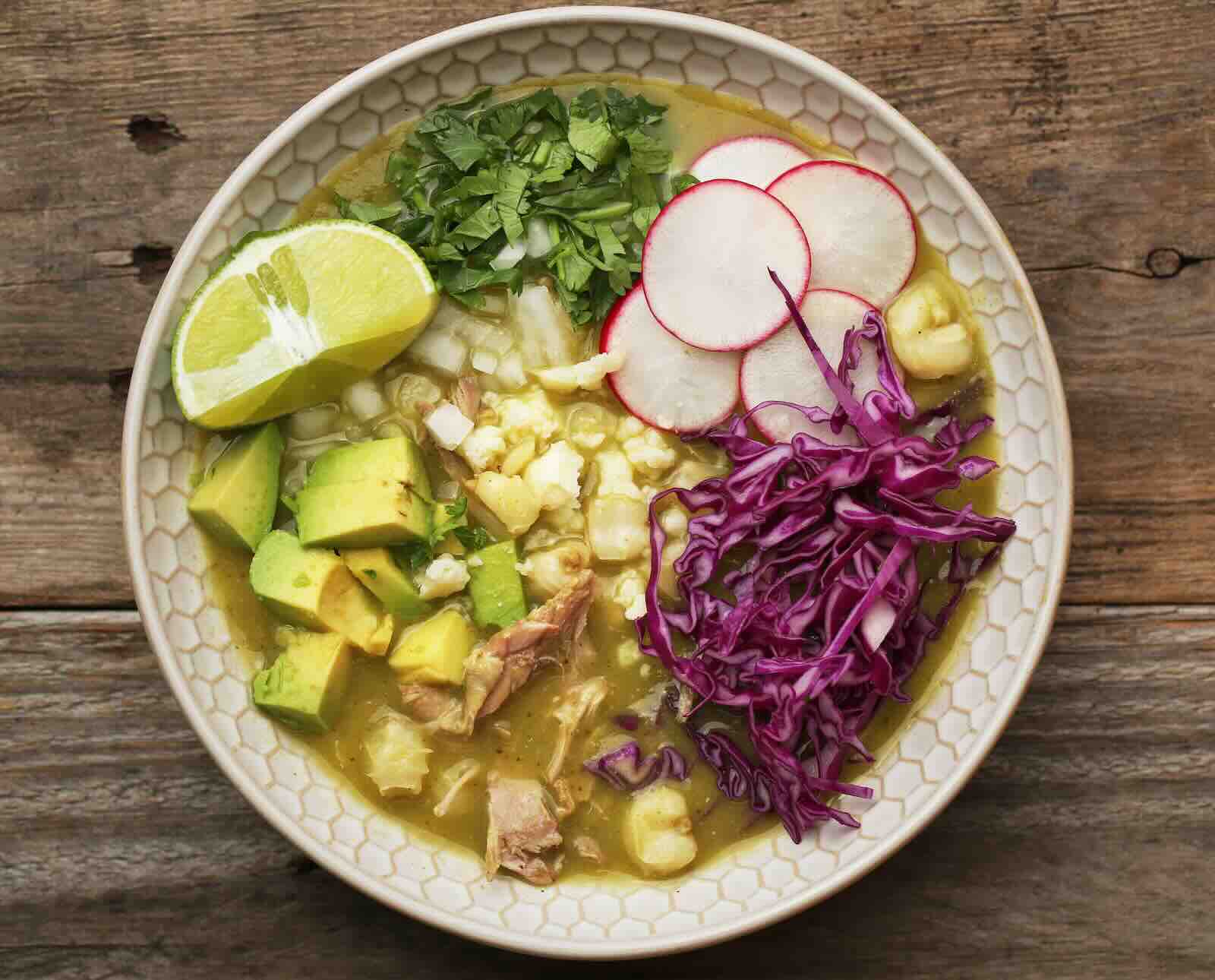 A white bowl of quail pozole topped with avocado, red cabbage, radish, cilantro, and lime wedges.