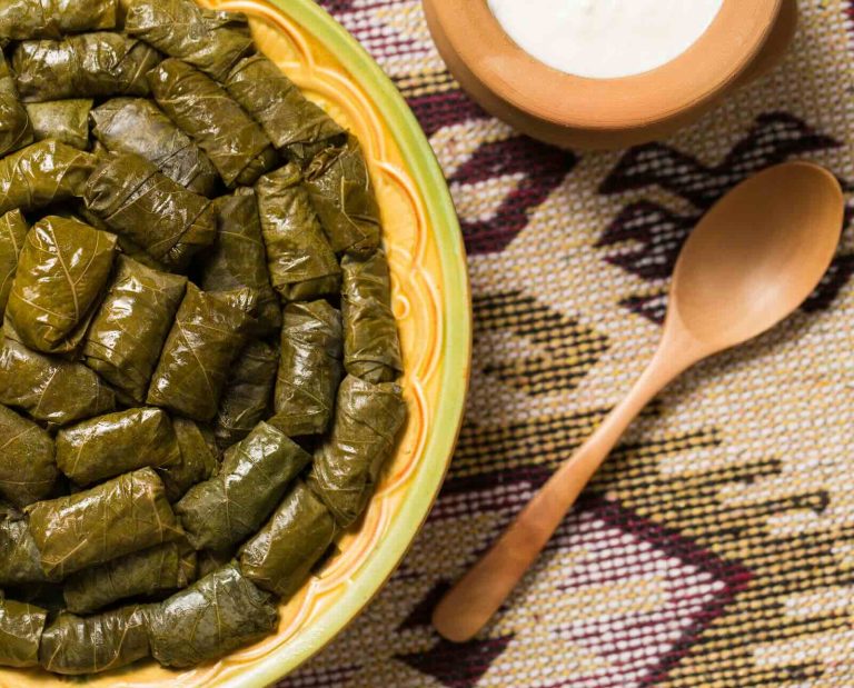 A plate of rolled dolmas served on a woven placemat with a wooden spoon and a bowl of white sauce