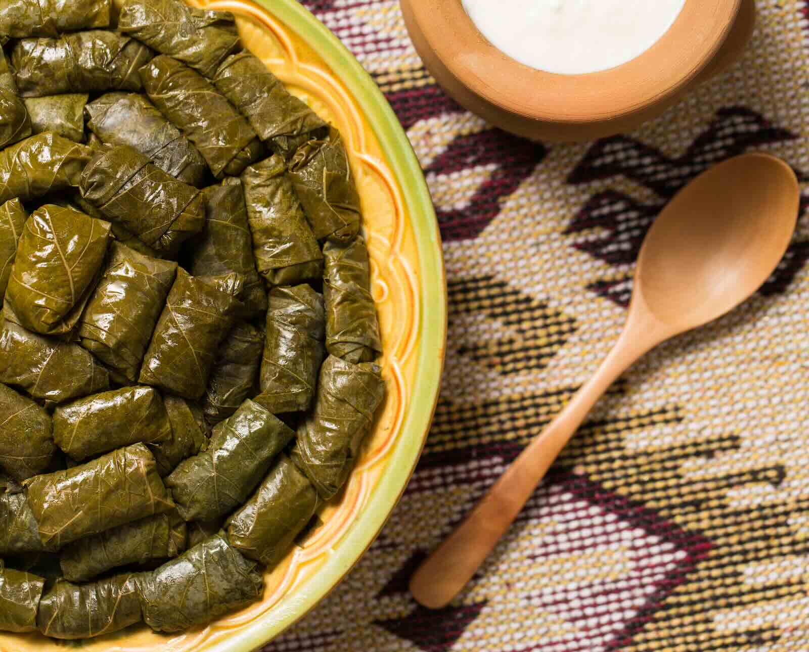 A plate of rolled dolmas served on a woven placemat with a wooden spoon and a bowl of white sauce