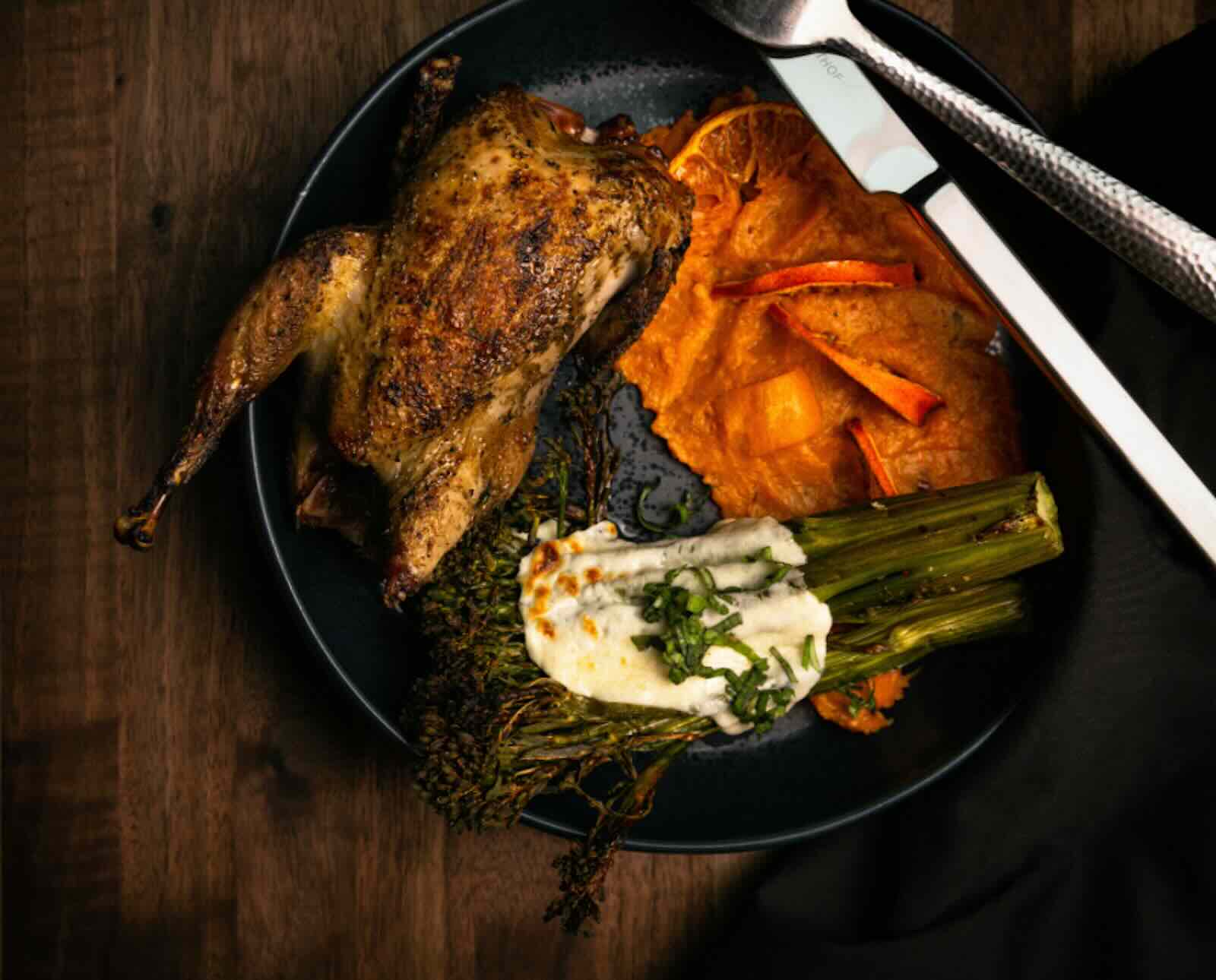 A whole roasted chukar, sweet potato puree, and broccoli are served on a plate with a fork and knife on a dark wooden background