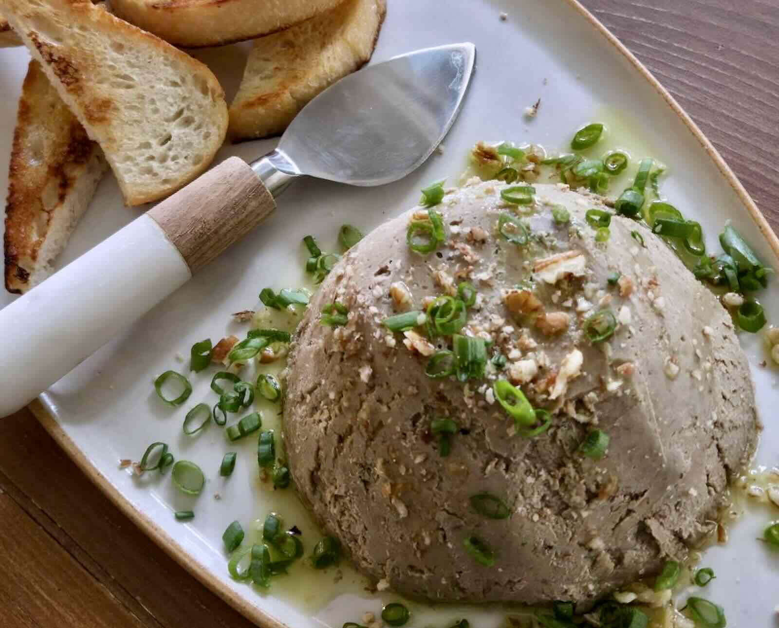 A mound of dove pate, topped with sliced green onions, is served on a white oval plate with toasted bread and a serving knife