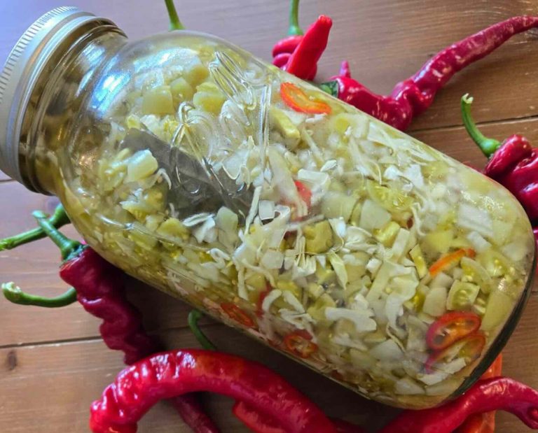 A mason jar filled with green tomato relish on a wooden background, surrounded with decorative red peppers