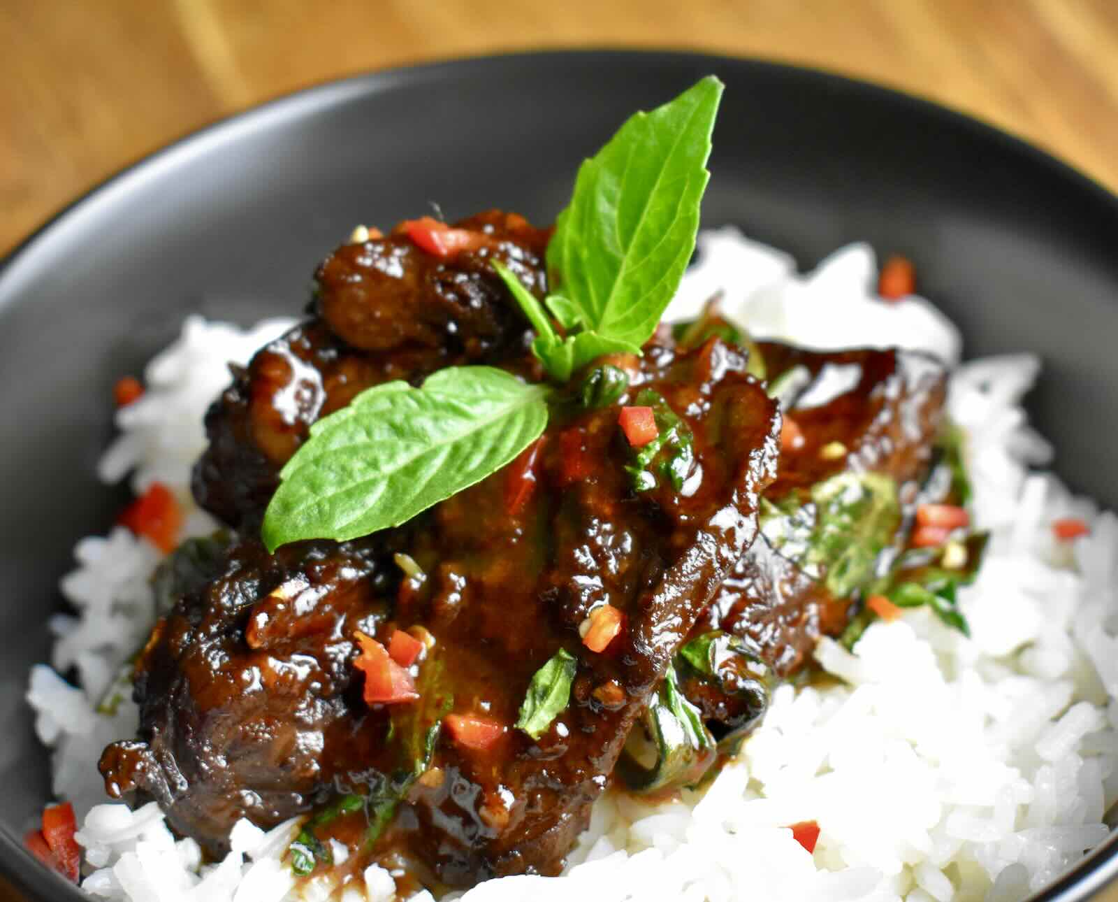 Roasted muskrat meat coated in a brown sauce, served atop white rice in a black bowl and garnished with Thai basil