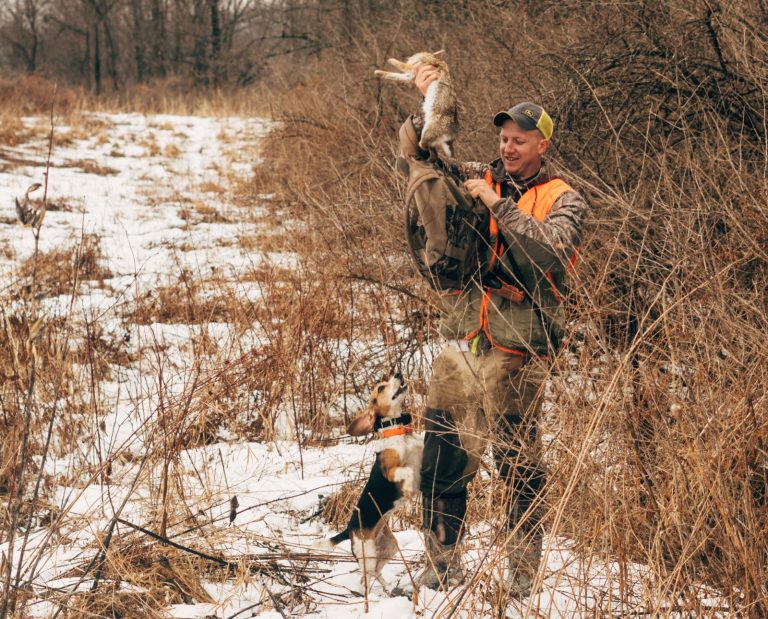 A rabbit hunter holds up a rabbit next to his excited beagle.