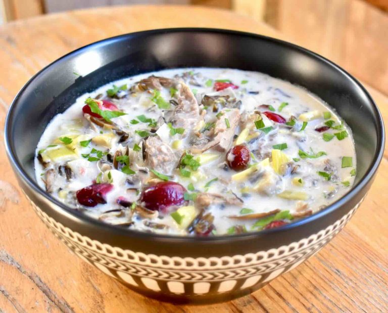 A patterned bowl on a wooden table filled with chowder made of shredded grouse meat, cranberries, and wild rice